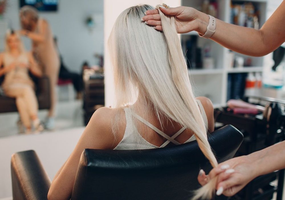 Hairdresser female making hair extensions to young woman with blonde hair in beauty salon. Professional hair extension salon